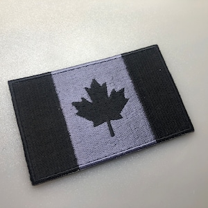 May include: A black and grey embroidered patch featuring the Canadian flag with a black maple leaf on a grey background.