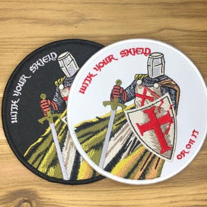 May include: Two round embroidered patches featuring a knight in armor holding a sword and shield with a red cross. One patch is black with white text, the other is white with red text. The text reads "WITH YOUR SHIELD OR ON IT".