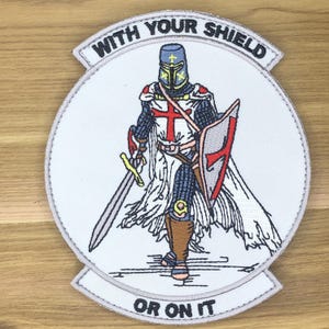 Puede incluir: Parche bordado con un caballero medieval con armadura completa, sosteniendo una espada y un escudo. El caballero tiene una cruz roja en el pecho y el escudo. El parche es blanco con el texto "WITH YOUR SHIELD" y "OR ON IT".