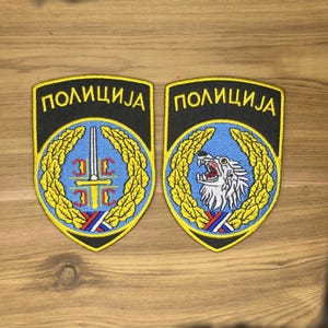 Pode incluir: Dois patches de polícia bordados. Cada patch tem um fundo preto com a palavra "ПОЛИЦИЈА" em amarelo. Um patch apresenta um design de espada e cruz, o outro um leão rugindo. Ambos têm uma coroa azul e dourada.