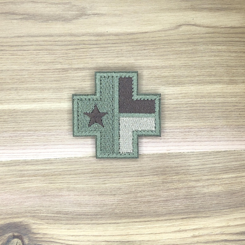 Puede incluir: Parche bordado en forma de cruz con una estrella y dise&ntilde;o geom&eacute;trico. El parche presenta tonos de verde, gris y blanco. La estrella es gris oscuro. El parche est&aacute; sobre una superficie de madera.