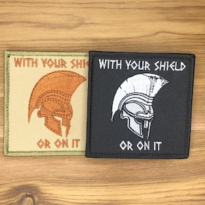 May include: Two square patches with embroidered text and imagery. One patch is tan with an orange Spartan helmet and the words "WITH YOUR SHIELD" and "OR ON IT." The other is black with a white helmet and the same text.