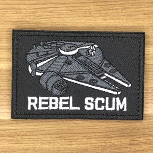 May include: A black rectangular patch with a grey and white spaceship design. The text "REBEL SCUM" is embroidered in white below the spaceship. The patch has a black border.