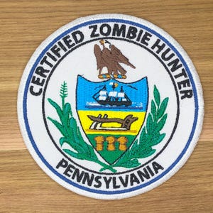 Könnte beinhalten: Ein runder Aufnäher mit weißem Hintergrund und blauem Rand. Der Aufnäher zeigt den Text "CERTIFIED ZOMBIE HUNTER" und "PENNSYLVANIA" in einem kreisförmigen Muster. Die Mitte zeigt ein farbenfrohes Emblem mit einem Adler und einem Schiff.