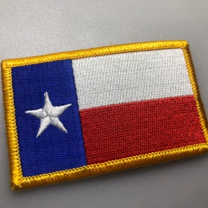 Puede incluir: Un parche bordado azul, blanco y rojo con una estrella blanca en la sección azul. El parche está rodeado por un borde dorado.