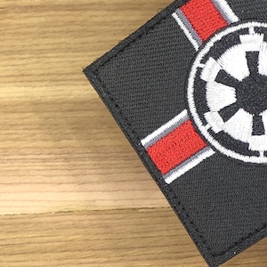 Może przedstawiać: Czarna kwadratowa naszywka z białym i czerwonym emblematem. Emblemat zawiera okrągły wzór z centralnym symbolem oraz czerwonymi i białymi paskami. Naszywka jest przeznaczona na odzież lub akcesoria.