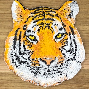 May include: Embroidered patch depicting a tiger's head. The patch is detailed with orange, black, white, and yellow. The tiger's face is central, with its eyes and stripes clearly visible. The patch is likely for clothing or accessories, and is a decorative item.