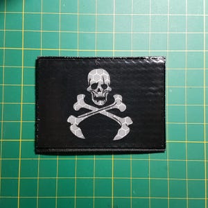 Könnte beinhalten: Ein schwarzer Patch mit einem weißen Totenkopf- und Knochenkreuz-Design. Der Totenkopf und das Knochenkreuz sind mit einem reflektierenden Material umrandet.