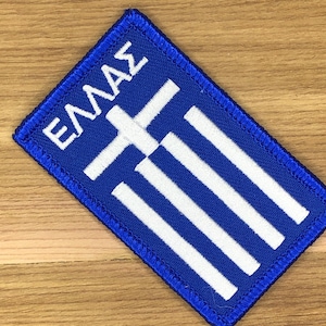 Puede incluir: Parche bordado azul y blanco con el diseño de la bandera griega. El parche tiene un borde azul y la palabra "ΕΛΛΑΣ" en letras blancas en la parte superior. El diseño incluye una cruz blanca y rayas blancas.