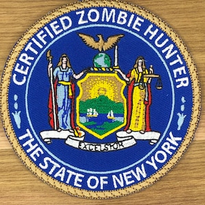 Peut inclure: Écusson brodé circulaire avec le texte "CERTIFIED ZOMBIE HUNTER" et "THE STATE OF NEW YORK". L'écusson présente le sceau de l'État de New York au centre, avec un fond bleu et une bordure dorée.