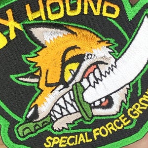 Könnte beinhalten: Gestickter Aufnäher mit einem Cartoon-Fuchs mit orange-weißem Fell, der eine grüne Klinge beißt. Der Aufnäher trägt den gelben Schriftzug "FOX HOUND" und "SPECIAL FORCE GROUP" mit grünem Rand.
