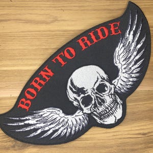 Può includere: Toppa nera con teschio bianco alato e scritta rossa "BORN TO RIDE". Il teschio è dettagliato, le ali sono bianche e piumate. La toppa ha la forma di un'ala.