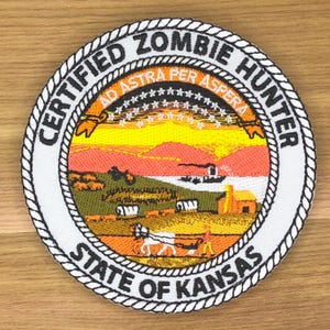 May include: Embroidered patch with a circular design featuring the text "CERTIFIED ZOMBIE HUNTER" and "STATE OF KANSAS". The patch has a detailed landscape scene with a sunset, fields, and a horse-drawn carriage. The border is white with a rope-like design.