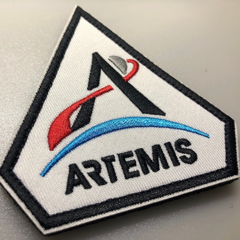 May include: A white embroidered patch with a black outline in the shape of a triangle. The patch features a stylised letter "A" with a red and blue curved line around it. The word "ARTEMIS" is embroidered in black below the letter "A".