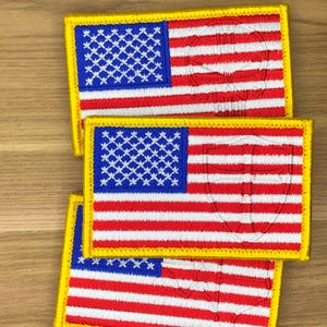 Puede incluir: Tres parches bordados con la bandera estadounidense. Cada parche tiene un campo azul con estrellas blancas, rayas rojas y blancas y un borde amarillo. Un diseño de escudo está sutilmente bordado sobre las rayas.