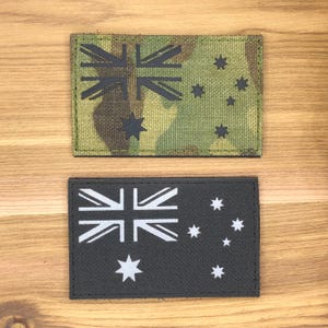 May include: Two rectangular patches displaying the Australian flag. One patch has a camouflage pattern with a black flag design. The other has a black background with a white flag design. Both are on a wooden surface.
