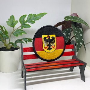 May include: A red, yellow and black embroidered patch featuring the German flag and eagle crest. The patch is displayed on a small wooden bench.