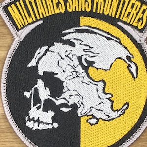 May include: Embroidered patch featuring a skull design with a map-like outline, split between black and white. The patch has a yellow and black background with the text "MILITAIRES SANS FRONTIERES" in yellow.