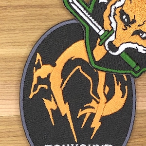 Puede incluir: Dos parches bordados. Uno es un óvalo negro con un diseño de criatura naranja parecida a un zorro y un rayo. El otro parche presenta una cabeza de zorro con una espada, acentos verdes y blancos. Ambos parches están sobre una superficie de madera.