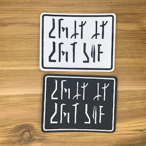 Puede incluir: Dos parches rectangulares, uno blanco y otro negro, cada uno con el mismo texto bordado en una fuente estilizada. El parche blanco tiene texto negro, y el parche negro tiene texto blanco. Ambos parches tienen un borde negro.