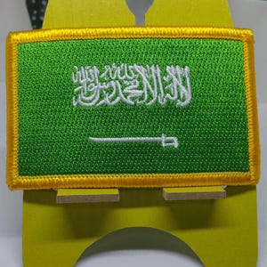 Puede incluir: Un parche verde bordado con una inscripción árabe blanca y una espada blanca. El parche tiene un borde amarillo y se muestra en un soporte amarillo.