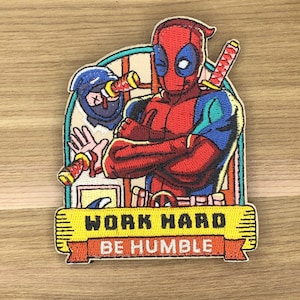 以下が含まれることがあります： 赤いと黒のスーツと剣を持った漫画のキャラクターが特徴の刺繍パッチ。パッチには、忍者のキャラクターと、黄色のバナーに「WORK HARD BE HUMBLE」というテキストが含まれています。