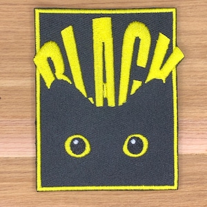 Puede incluir: Parche bordado con un diseño de gato estilizado. El parche rectangular tiene un fondo gris oscuro y un borde amarillo. La palabra "BLACK" está bordada en amarillo en la parte superior, con dos ojos amarillos debajo.