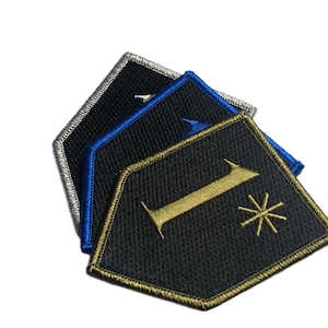 Pode incluir: Três patches bordados em forma de diamante. Os patches são pretos, azuis e prateados. O patch preto tem um "1" dourado e um desenho de estrela. Os patches são provavelmente para roupas ou acessórios.