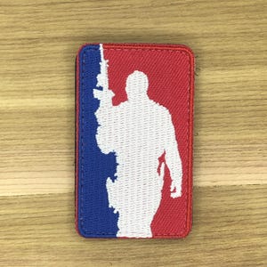 Könnte beinhalten: Rechteckiger Aufnäher mit rotem und blauem Hintergrund, der eine weiße Silhouette einer Person mit einem Gewehr zeigt. Der Aufnäher hat einen genähten Rand und ist wahrscheinlich ein Emblem oder eine Insignie.