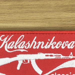 Può includere: Una toppa rossa con testo ricamato bianco "Kalashnikova" sopra un'immagine ricamata bianca di un fucile d'assalto AK-47. La toppa è rettangolare e ha una superficie strutturata.