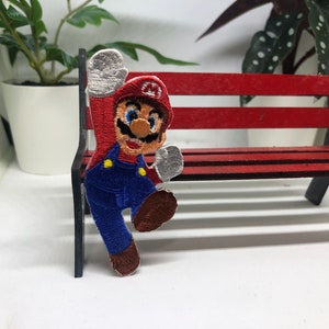 Könnte beinhalten: Gestickter Bügel-Patch mit einer Comicfigur, die einen roten Hut, eine blaue Latzhose und weiße Handschuhe trägt. Die Figur ist in einer Sprungpose mit einem erhobenen Arm.