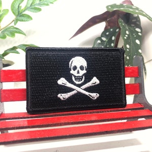 May include: A black fabric patch with a white embroidered skull and crossbones design. The patch is sitting on a red wooden bench.