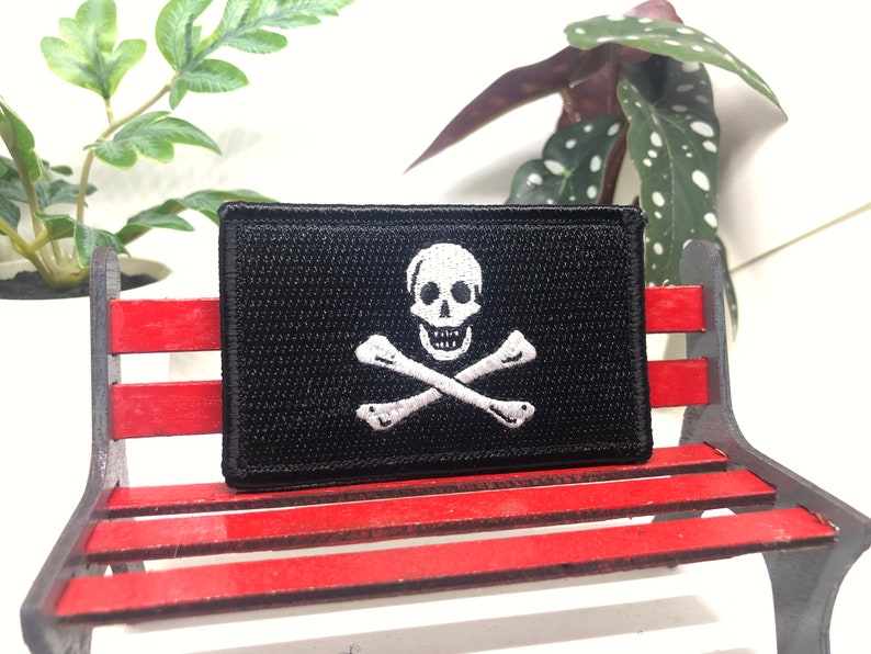 May include: A black fabric patch with a white embroidered skull and crossbones design. The patch is sitting on a red wooden bench.