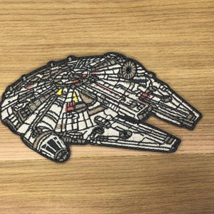 Puede incluir: Parche bordado con la nave espacial Millennium Falcon. El parche tiene un diseño detallado con detalles en blanco, gris, negro, amarillo y rojo. El parche tiene forma ovalada con tres secciones que sobresalen.