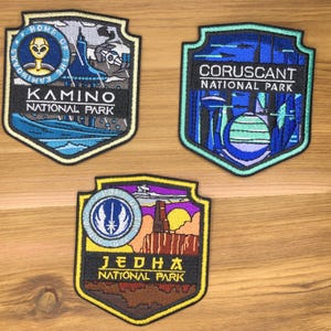 Pode incluir: Três patches bordados com desenhos de escudo. Um patch diz "Kamino National Park" com um alienígena e uma cena aquática. Outro diz "Coruscant National Park" com uma paisagem urbana. O terceiro diz "JEDHA National Park" com uma cena desértica.