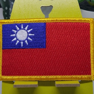 Puede incluir: Un parche bordado con la bandera de Taiwán. El parche es rojo, blanco y azul con un borde amarillo.