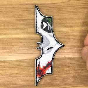 Op de afbeelding: Geborduurde patch in de vorm van een vleermuissymbool, met een op de Joker geïnspireerd ontwerp. De patch heeft een witte achtergrond met groene, zwarte, rode en grijze details. De patch ligt op een houten oppervlak.