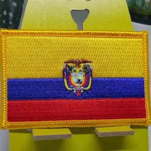 May include: A yellow, blue, and red embroidered patch with the coat of arms of Ecuador in the center. The patch has a yellow border.