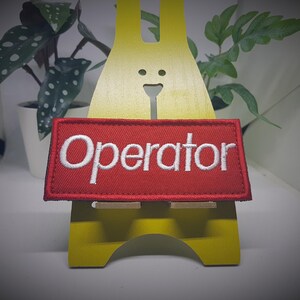 Może przedstawiać: Czerwona naszywka haftowana z białym napisem "Operator".