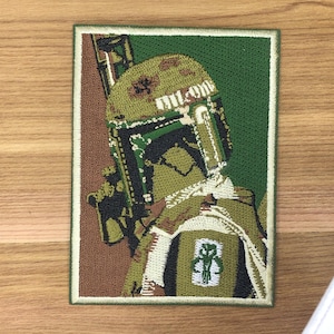 May include: Embroidered patch depicting a character from a science fiction film. The patch features a green, brown, and white colour scheme. The character is wearing a helmet and armour, and holding a weapon. The patch is rectangular with a green border.