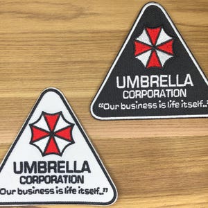 Puede incluir: Dos parches triangulares con el texto "UMBRELLA CORPORATION" y la cita "Our business is life itself." Los parches presentan un emblema rojo y blanco. Uno es negro con texto blanco, y el otro es blanco con texto negro.