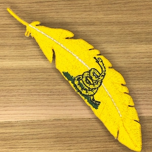 May include: Embroidered yellow feather patch featuring a coiled snake and the words "Don't Tread on Me" in black and green. The patch has a white stitched spine and is set against a wooden background.