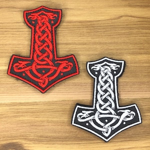 May include: Two embroidered patches in the shape of Thor's hammer, a symbol of Norse mythology. One patch is black with red detailing, the other is black with white detailing. Both feature intricate knotwork designs.