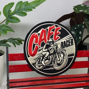 May include: A black and white embroidered patch with red lettering that says "Cafe Racer". The patch features a rider on a motorcycle.