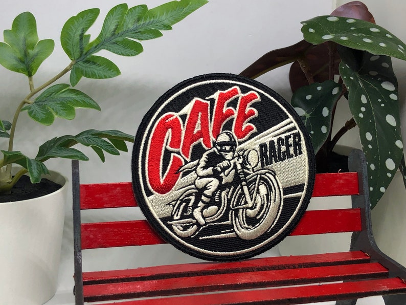 May include: A black and white embroidered patch with red lettering that says "Cafe Racer". The patch features a rider on a motorcycle.