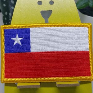 May include: A red, white, and blue Chilean flag patch with a white star in the blue corner. The patch is surrounded by a gold border and is displayed on a yellow stand.