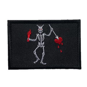 May include: A black embroidered patch depicting a white skeleton holding a flaming torch and a spear through a red heart. The skeleton has red eyes, set against a black background.