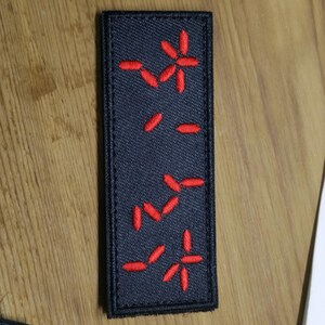 Puede incluir: Un parche rectangular negro con dise&ntilde;os bordados en rojo. El parche presenta varias formas rojas estilizadas en forma de flor o estrella y l&iacute;neas cortas. El parche es probablemente para ropa o accesorios.
