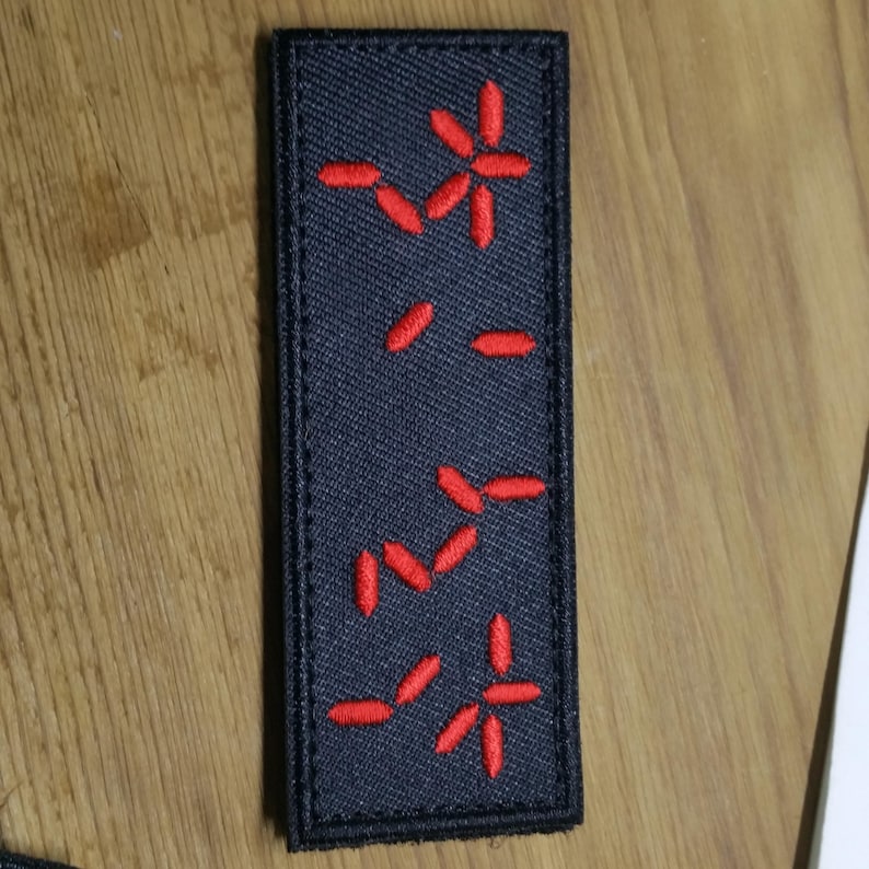 Puede incluir: Un parche rectangular negro con dise&ntilde;os bordados en rojo. El parche presenta varias formas rojas estilizadas en forma de flor o estrella y l&iacute;neas cortas. El parche es probablemente para ropa o accesorios.