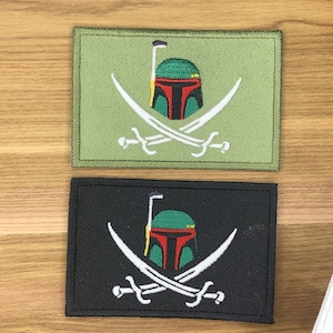 May include: Two rectangular patches featuring a helmet design with crossed swords. One patch is green with a red, green, and yellow helmet and white swords. The other patch is black with the same helmet and white swords.
